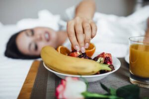 A woman reaching for fresh fruit on a tray, resting in bed with a glass of juice and a rose nearby. Soft, cozy atmosphere.