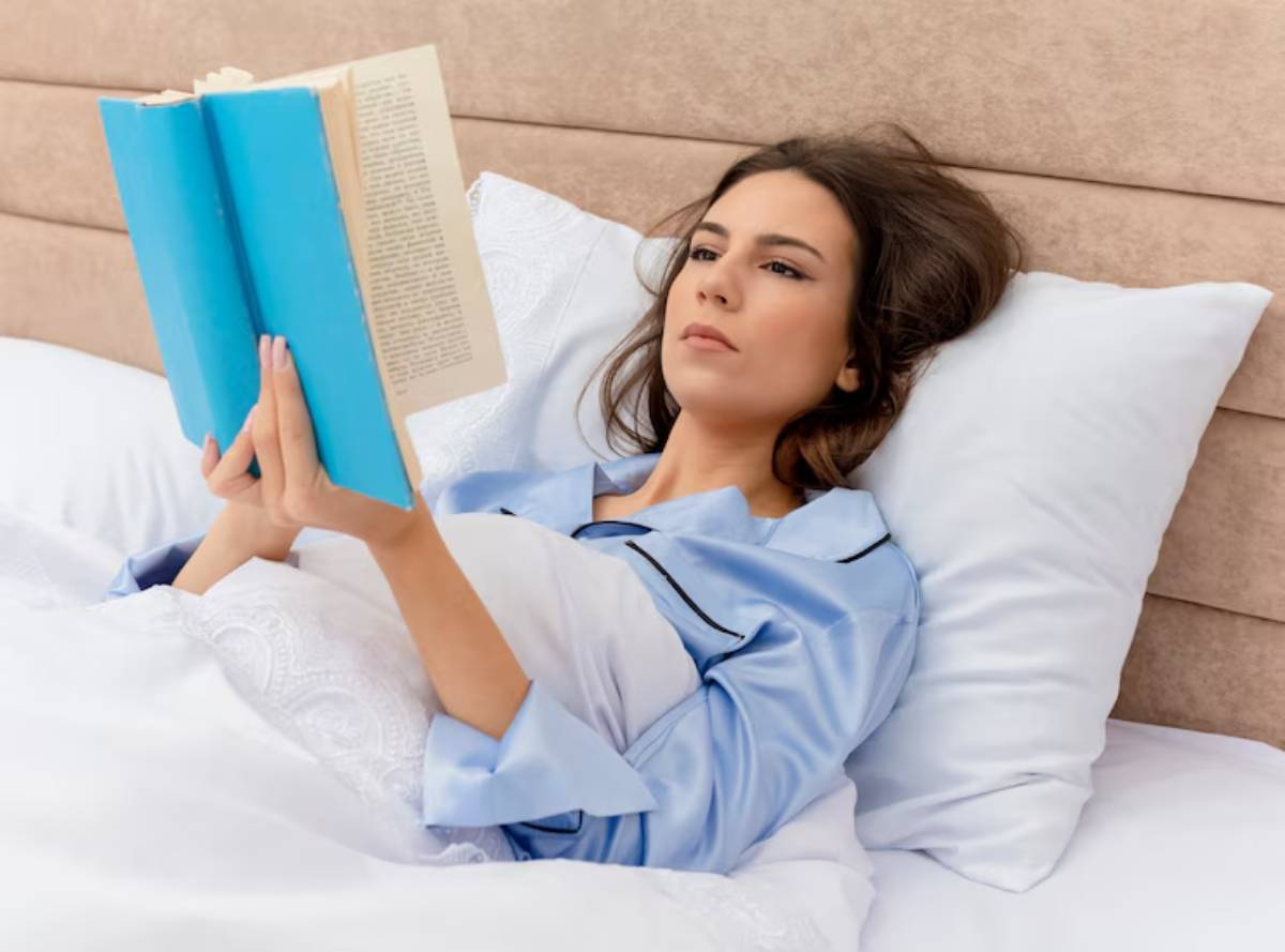 A person in blue pajamas lies in bed, comfortably reading a large blue book, surrounded by fluffy pillows and soft bedding.