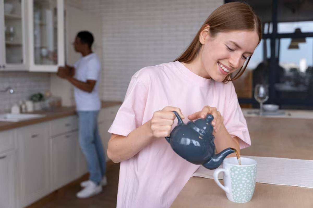 A person in a pink shirt pours tea from a dark kettle into a light blue polka-dotted mug in a cozy, modern kitchen.
