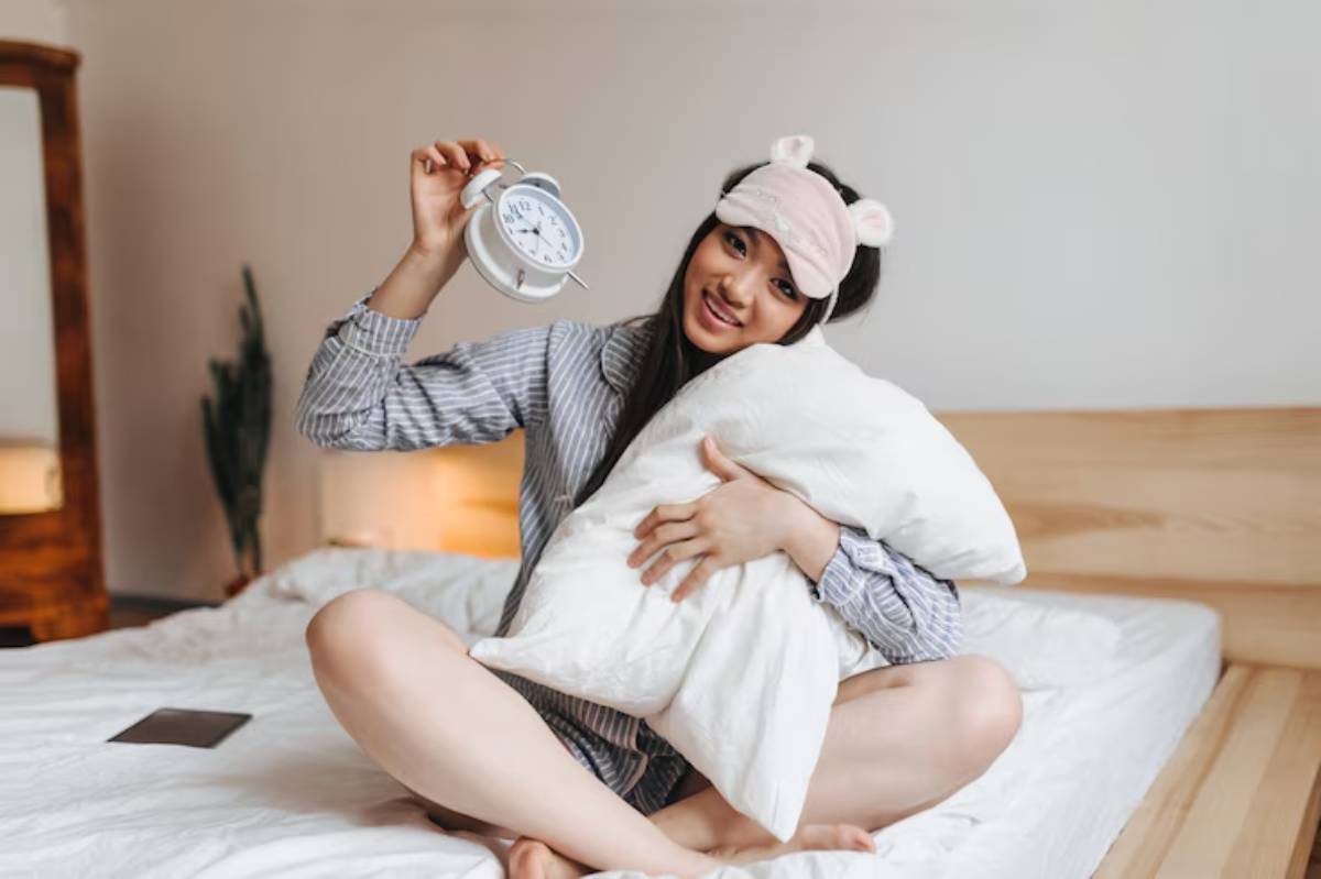 A person in pajamas holds a clock and snuggles a pillow while sitting on a bed in a cozy, softly lit bedroom setting.