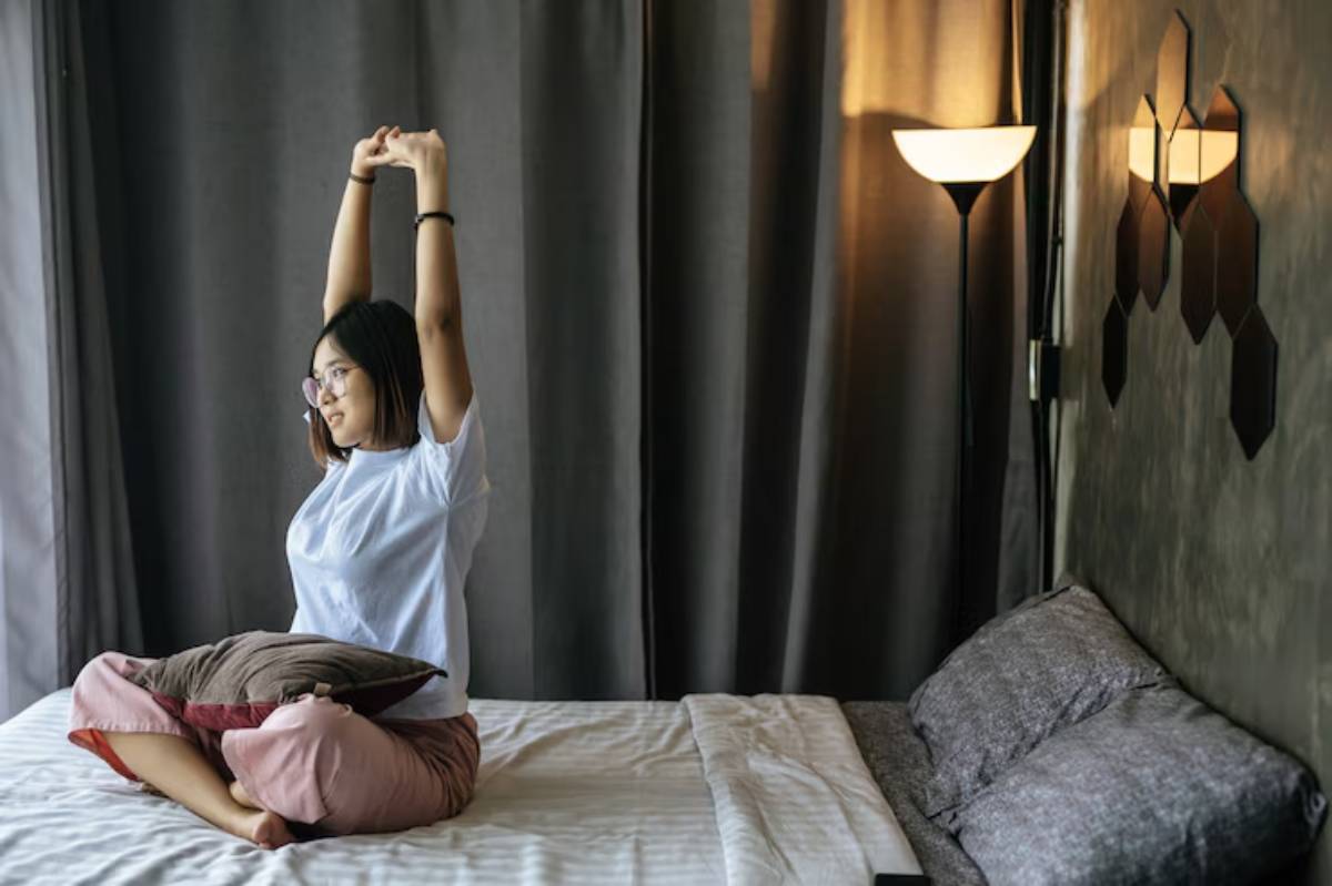 A person stretches while seated with crossed legs on a bed, surrounded by curtains and a lamp, creating a tranquil indoor atmosphere.