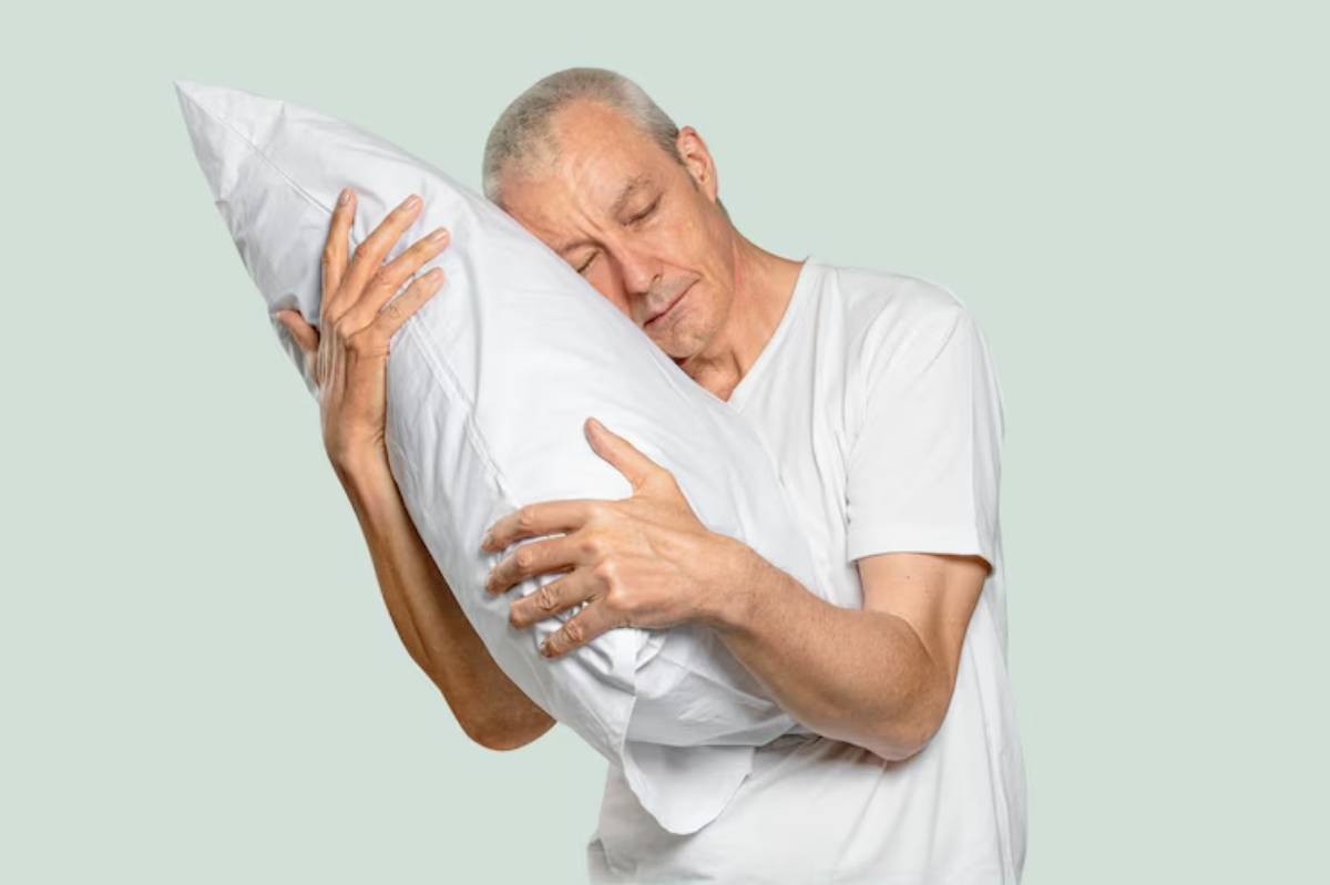 An elderly person in a white t-shirt tenderly hugs a fluffy white pillow against a soft green background.