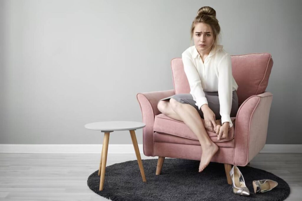A woman in a white blouse and gray skirt sits on a pink armchair, removing a shoe, with a small round table nearby.
