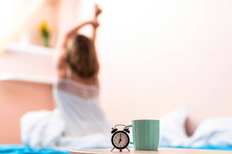 A woman stretches in bed, with a vintage alarm clock and a mint green mug in focus on the bedside table. Soft morning light filters in.