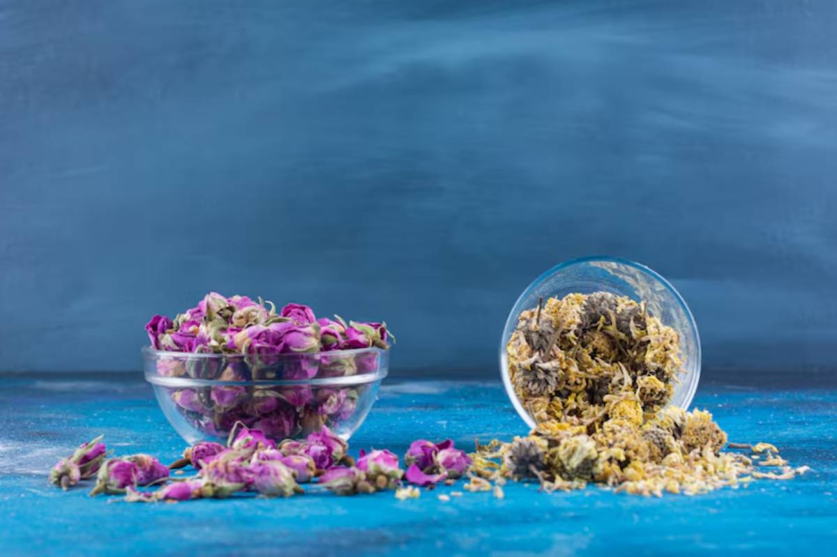 A bowl of dried purple rosebuds next to a glass dish spilling yellow and gray herbal mixture, set against a blue background.