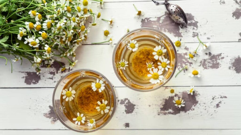 Two glasses of chamomile tea adorned with floating flowers, surrounded by fresh chamomile blooms on a rustic wooden table.