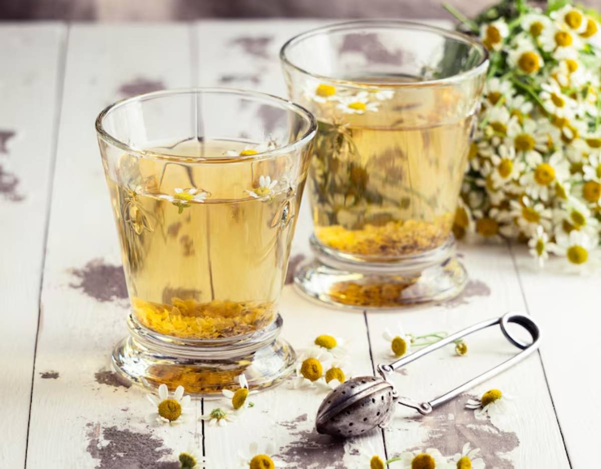 Two glasses of chamomile tea with flowers, surrounded by fresh chamomile blossoms and a tea infuser on a rustic wooden table.
