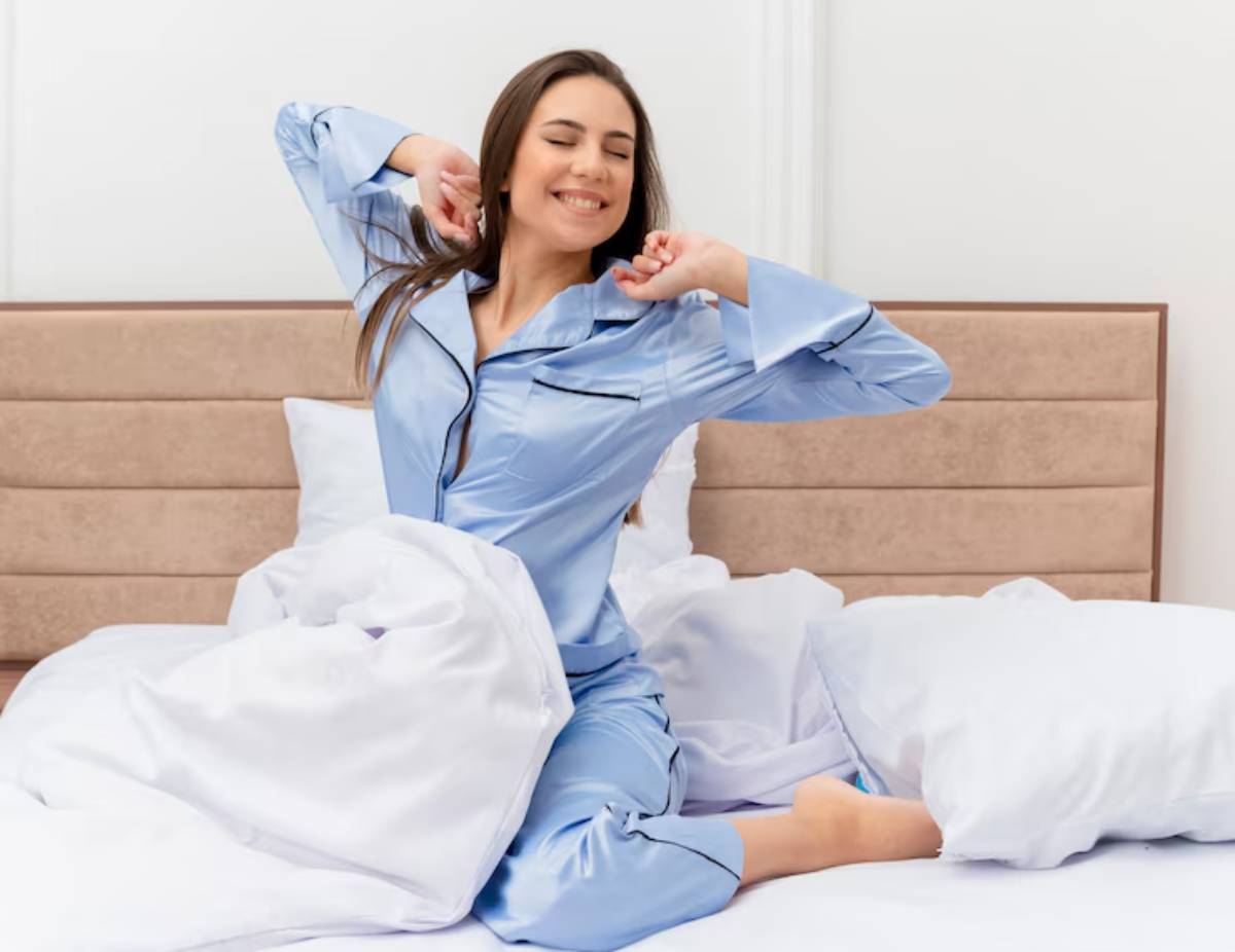 A person in blue pajamas stretches while sitting on a bed surrounded by fluffy white pillows and a cozy blanket.