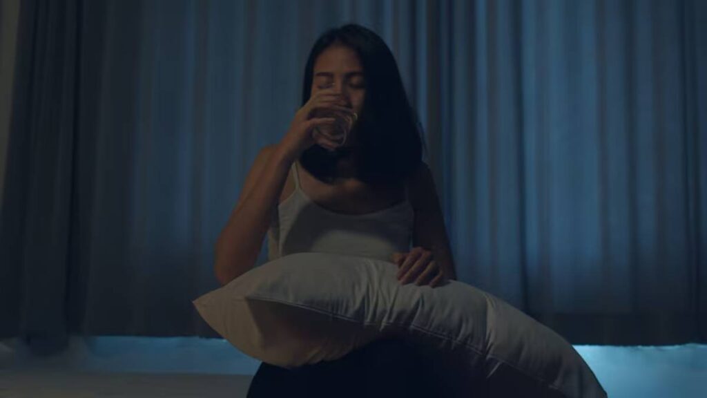 A woman sits on a bed holding a pillow, surrounded by dim light and soft curtain drapes, exuding a sense of contemplation or introspection.