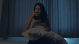 A woman sits on a bed holding a pillow, surrounded by dim light and soft curtain drapes, exuding a sense of contemplation or introspection.