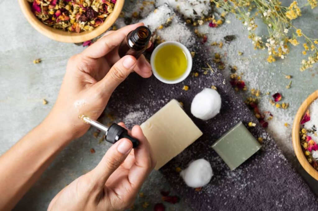 A person's hands apply essential oil from a dropper, surrounded by natural ingredients like flowers, soap, and cotton balls on a textured surface.