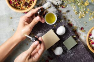A person's hands apply essential oil from a dropper, surrounded by natural ingredients like flowers, soap, and cotton balls on a textured surface.