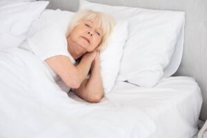 An elderly person lies on a bed, wrapped in white blankets, resting their head on pillows with a serene expression.