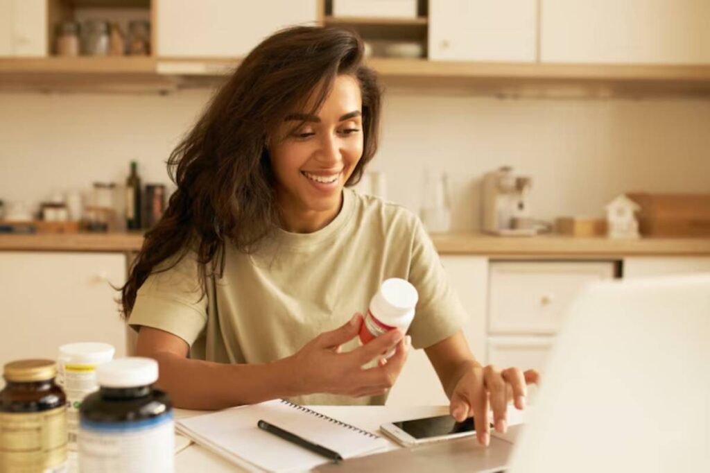 A person in a light-colored shirt examines a vitamin bottle while seated at a kitchen table with various supplements and a laptop.