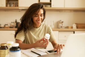 A person in a light-colored shirt examines a vitamin bottle while seated at a kitchen table with various supplements and a laptop.