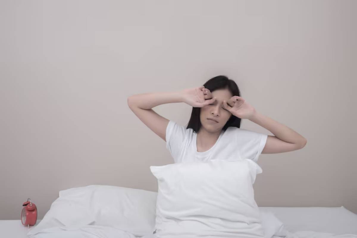 A person in a white t-shirt sits up in bed, resting against white pillows with a red alarm clock visible on the bedside table.