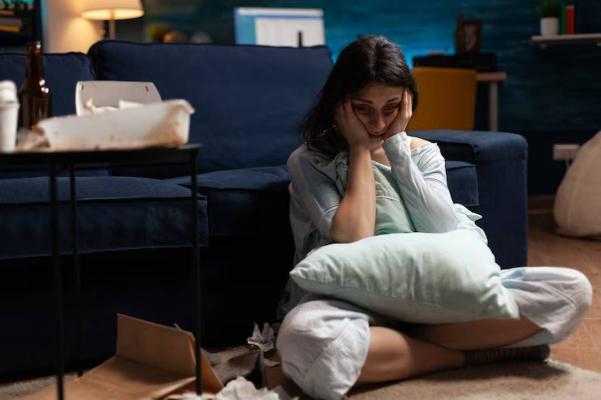 A person sits on a living room floor, hugging a pillow, surrounded by discarded containers and a messy environment.