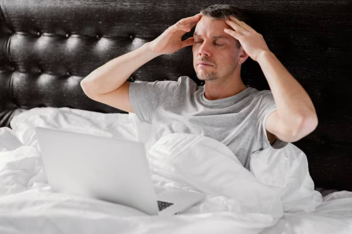 A person sitting in bed, surrounded by crumpled white sheets, appears stressed while looking at a laptop.