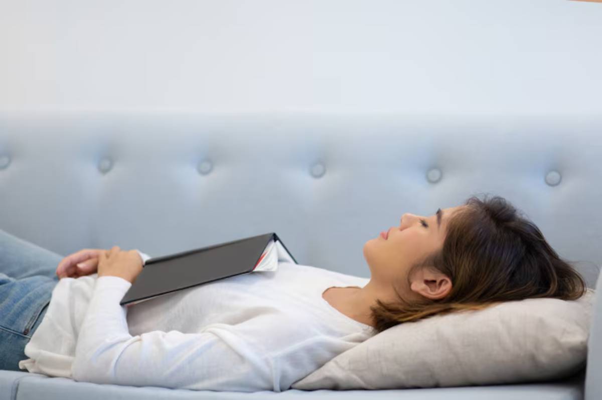 A person lies relaxed on a light gray couch, holding a black book on their chest, surrounded by soft pillows.
