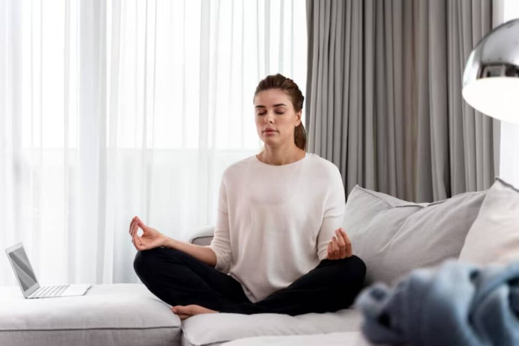 A person in a white sweater sits cross-legged on a sofa, meditating in a bright, airy room with sheer curtains and a laptop nearby.