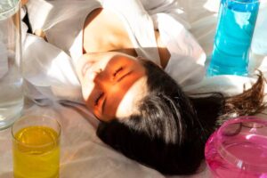 A person lies on a light surface surrounded by colorful drinks: a clear glass, a pink bowl, a bright yellow drink, and a blue liquid.