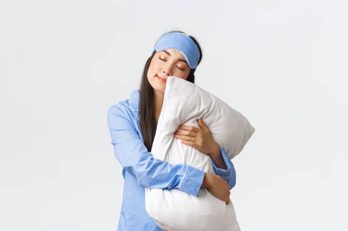 A woman in blue polka-dot pajamas hugs a large white pillow against a plain light background. She looks cozy and relaxed.
