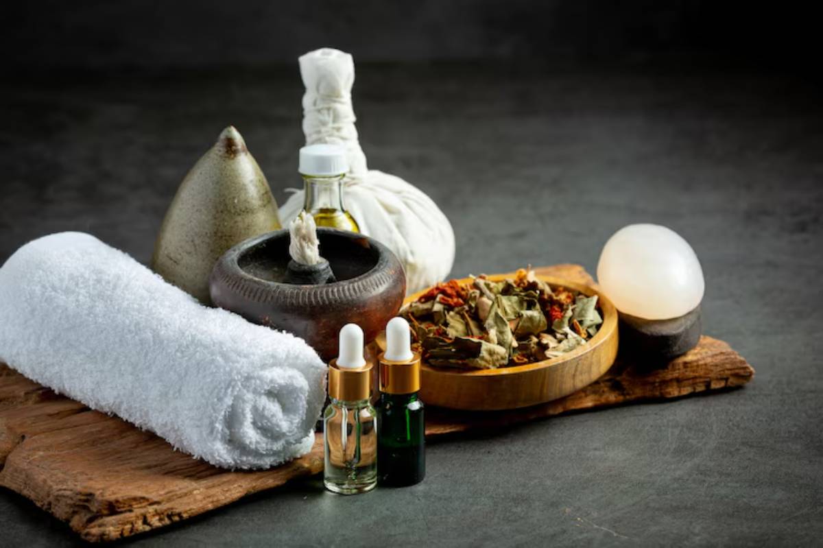 A collection of spa items: rolled towel, essential oil bottles, herbal blend in a bowl, and a stone, arranged on a wooden surface.