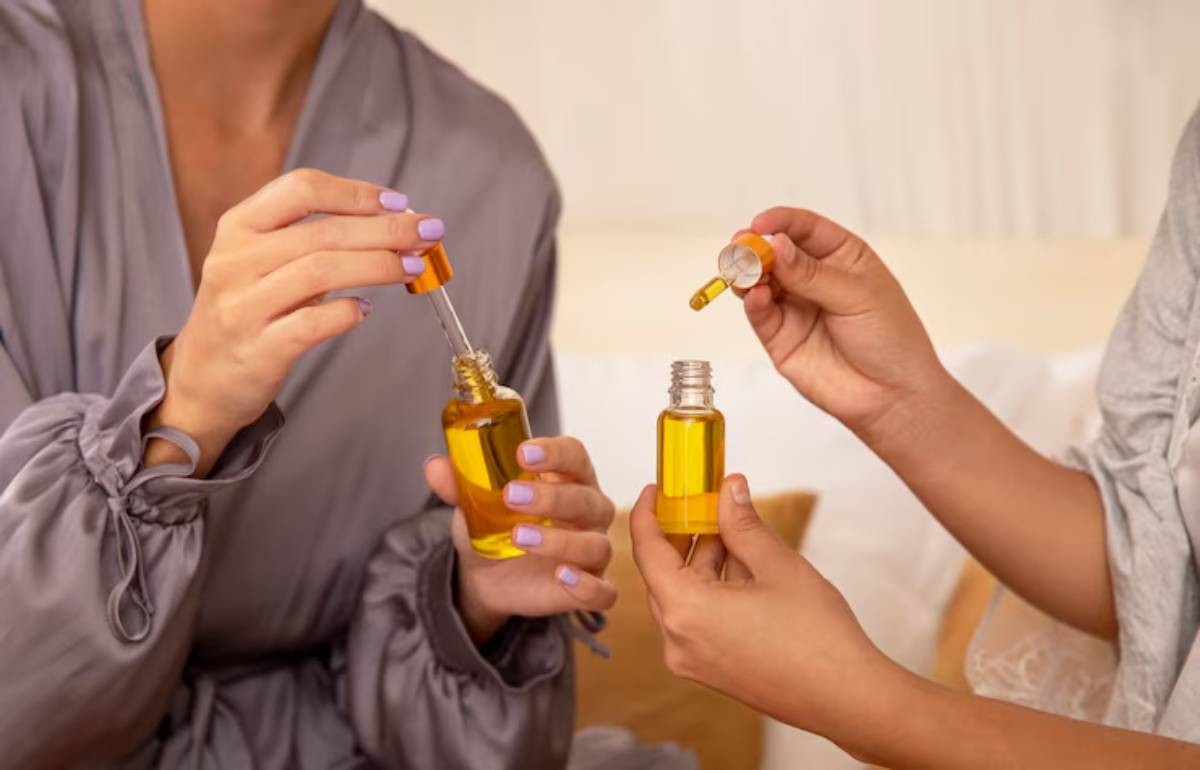 Two women in robes share beauty oil, one holding a dropper above a bottle while the other prepares to add to another bottle.