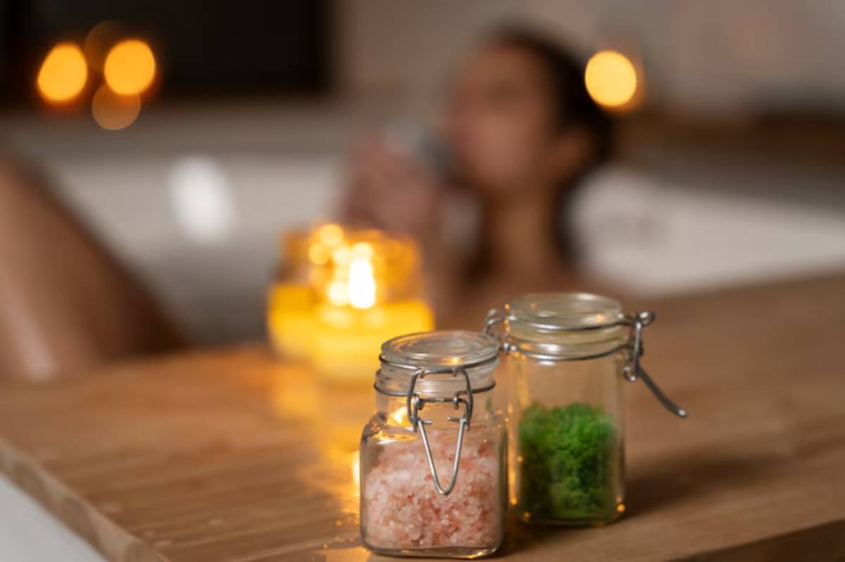 Two glass jars filled with pink bath salt and green bath additive sit on a wooden surface, glowing candles softly illuminate the background.
