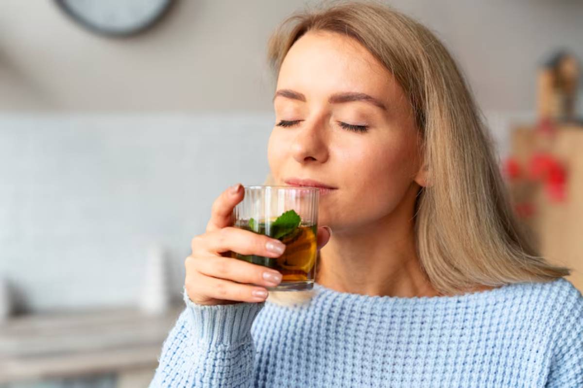 A woman in a light blue sweater holds a glass of amber liquid, sitting in a bright, cozy kitchen. A clock is visible in the background.
