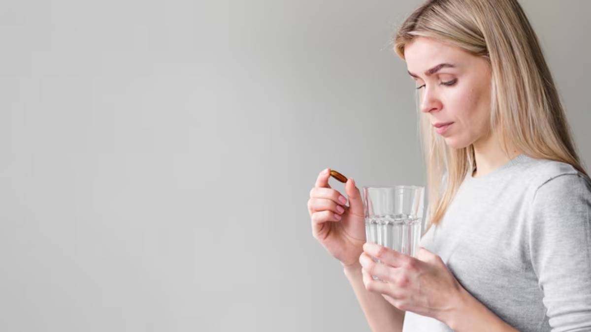 A person in a gray sweater holds a glass of water in one hand and a brown capsule in the other, preparing to take the supplement.