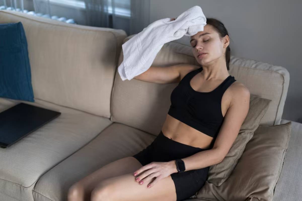 A woman in a black sports outfit sits on a couch, wiping her forehead with a towel, looking relaxed after a workout. 