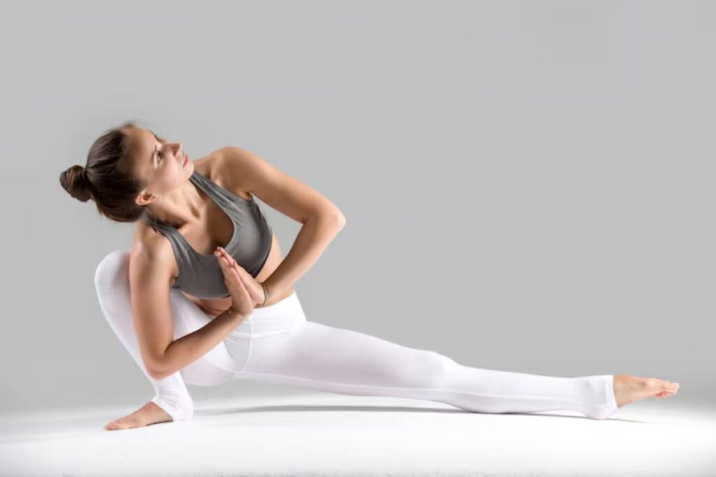 A woman performing a yoga pose with a graceful stance, dressed in a gray top and white leggings, on a light gray background.