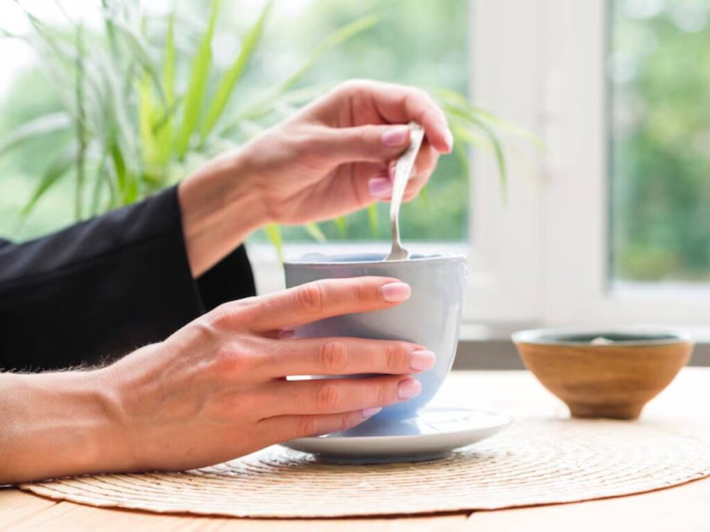 A hand stirs a light blue cup of tea or coffee with a spoon, sitting on a table with a woven placemat and a green plant in the background.