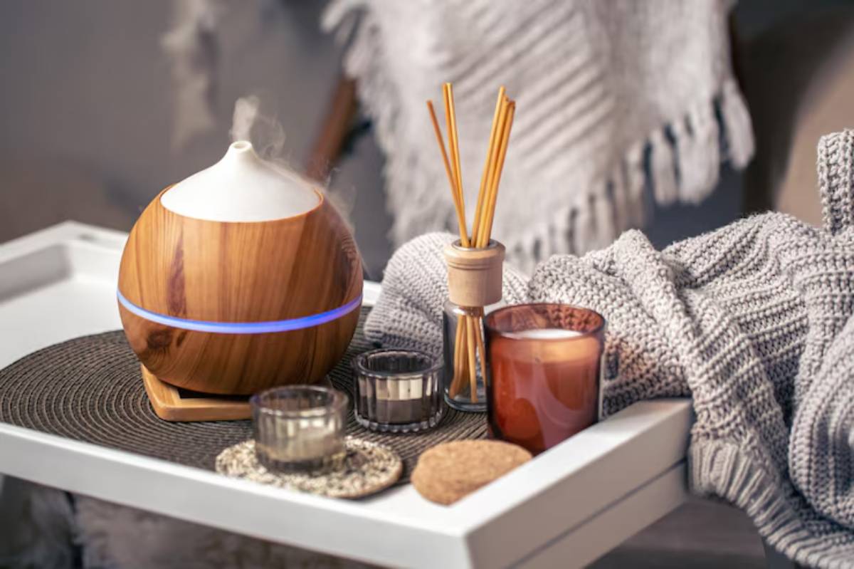 A wooden diffuser emits steam beside candles and decorative items on a cozy tray, with a soft gray blanket in the background.