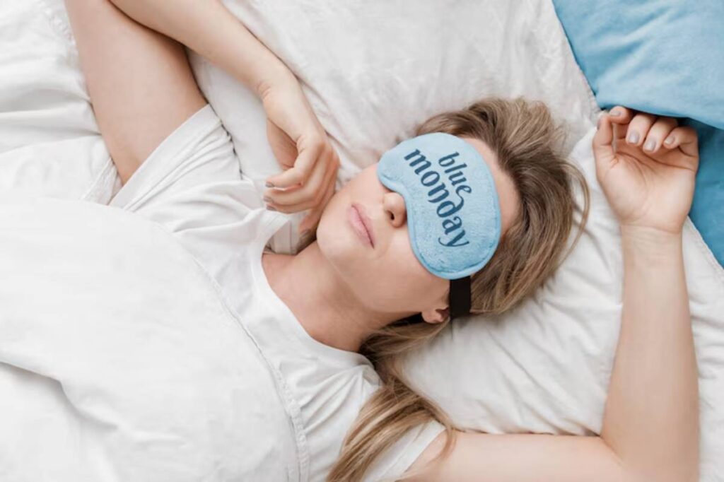 A person relaxing on a bed with white sheets, wearing a light blue sleep mask, with arms relaxed beside them.