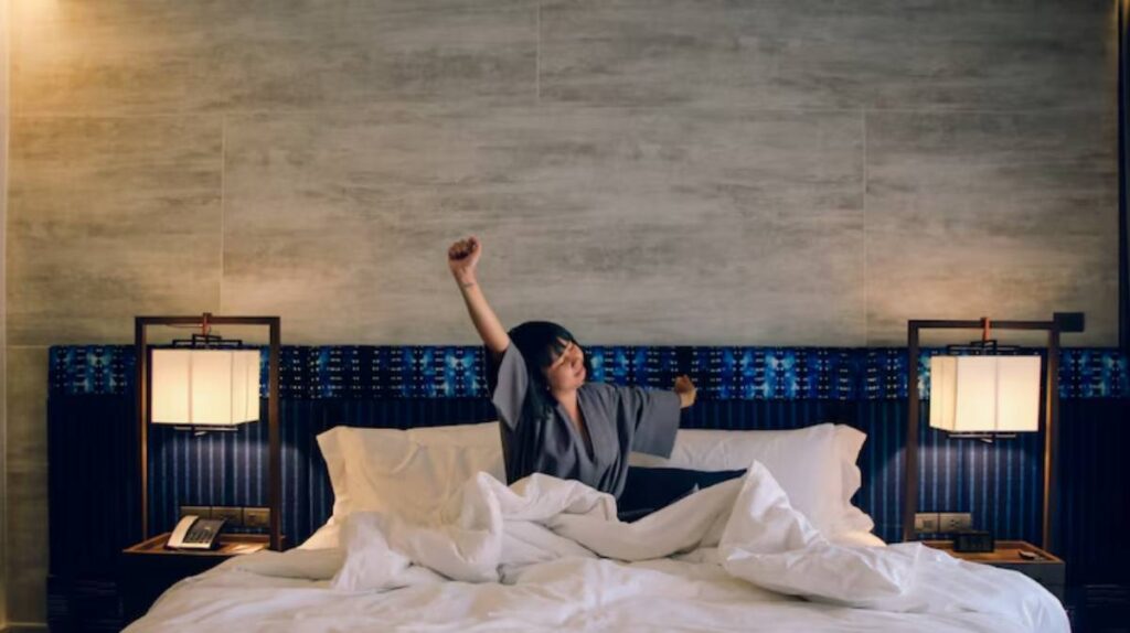 A person wearing a gray robe sits on a bed, stretching with arms raised, surrounded by cozy white bedding and modern room decor.