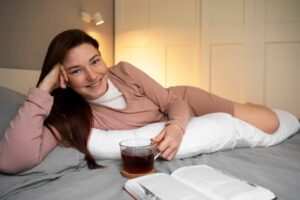 A woman relaxes on a bed, holding a cup of tea, with a book and soft pillows nearby, in a cozy, well-lit room.
