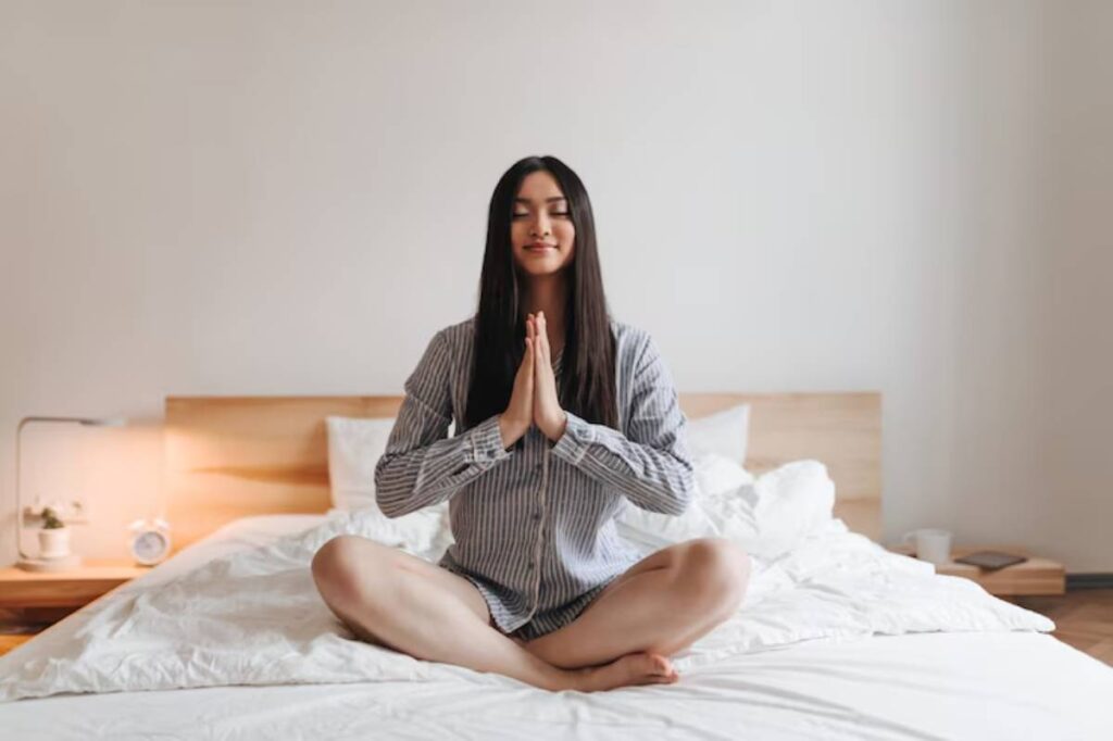 A person sits cross-legged on a bed, dressed in a striped shirt, with hands in a prayer position, exuding calm and mindfulness.