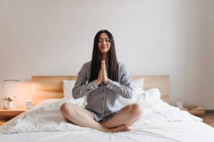 A person sits cross-legged on a bed, dressed in a striped shirt, with hands in a prayer position, exuding calm and mindfulness.
