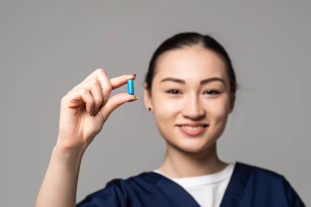 A medical professional in scrubs holds a blue capsule in their hand against a neutral gray background.