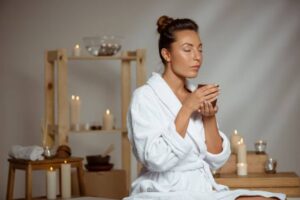 A woman in a white bathrobe holds a cup, surrounded by candles and wooden decorations, creating a serene and relaxing atmosphere.