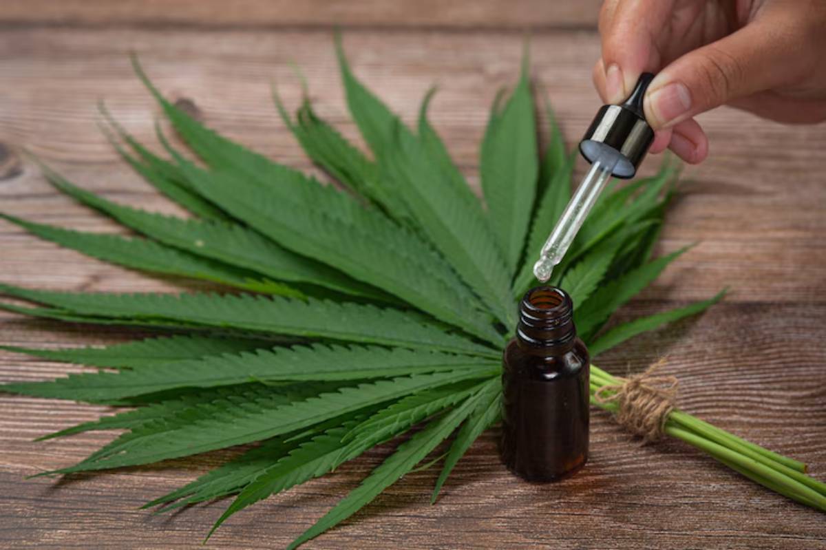 A hand holds a dropper above a dark bottle, surrounded by green cannabis leaves on a wooden surface.