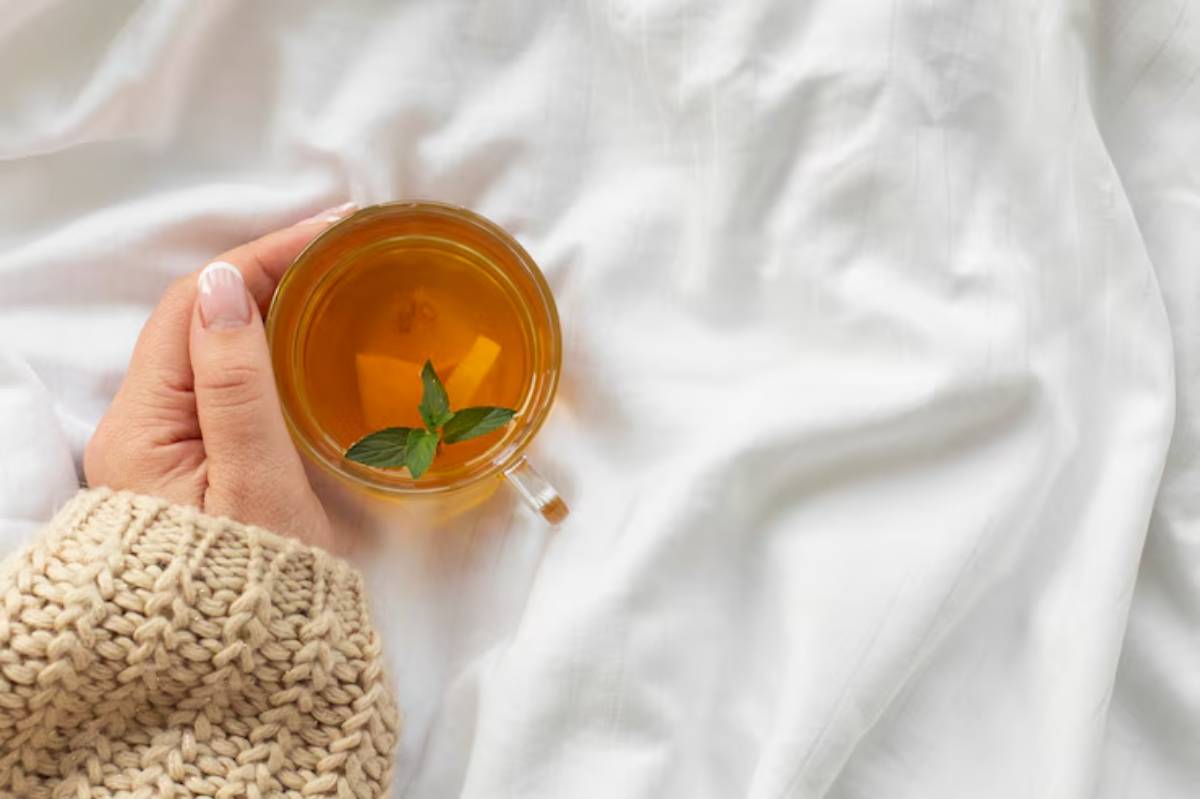 A person holds a glass of herbal tea with a mint sprig, resting on a soft, white blanket, wearing a cozy knitted sweater.