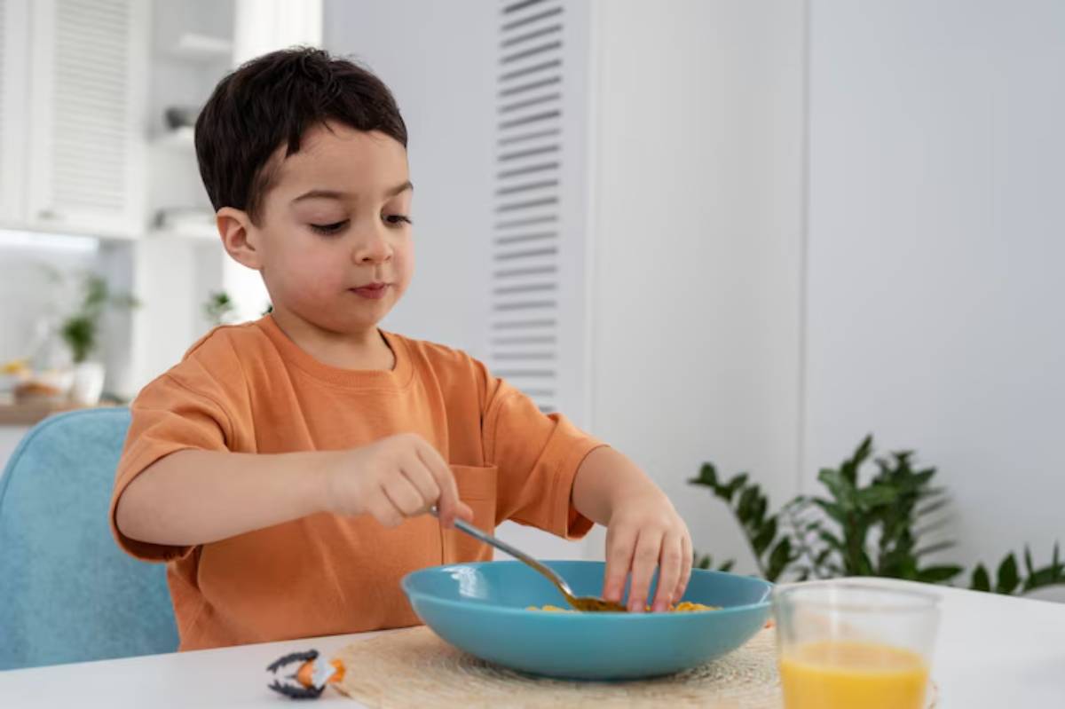 A child in an orange shirt eats from a blue bowl, with a glass of orange juice on the table and greenery in the background.
