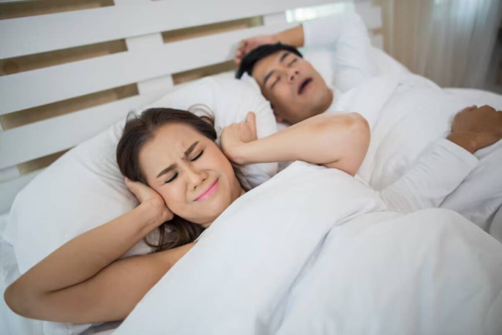 A couple relaxes in bed, cozy under white blankets, with soft morning light filtering through curtains.