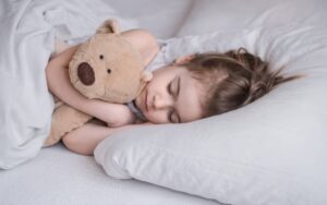A young girl peacefully sleeps on a white bed, cuddling a soft teddy bear, with her eyes closed and hair gently tousled.
