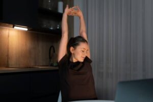 A person stretches their arms overhead while seated at a table in a dimly lit kitchen setting, creating a relaxed atmosphere.
