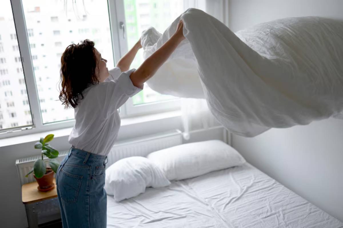 A woman in a white shirt and jeans lifts a duvet over a neatly made bed, sunlight streaming through large windows.