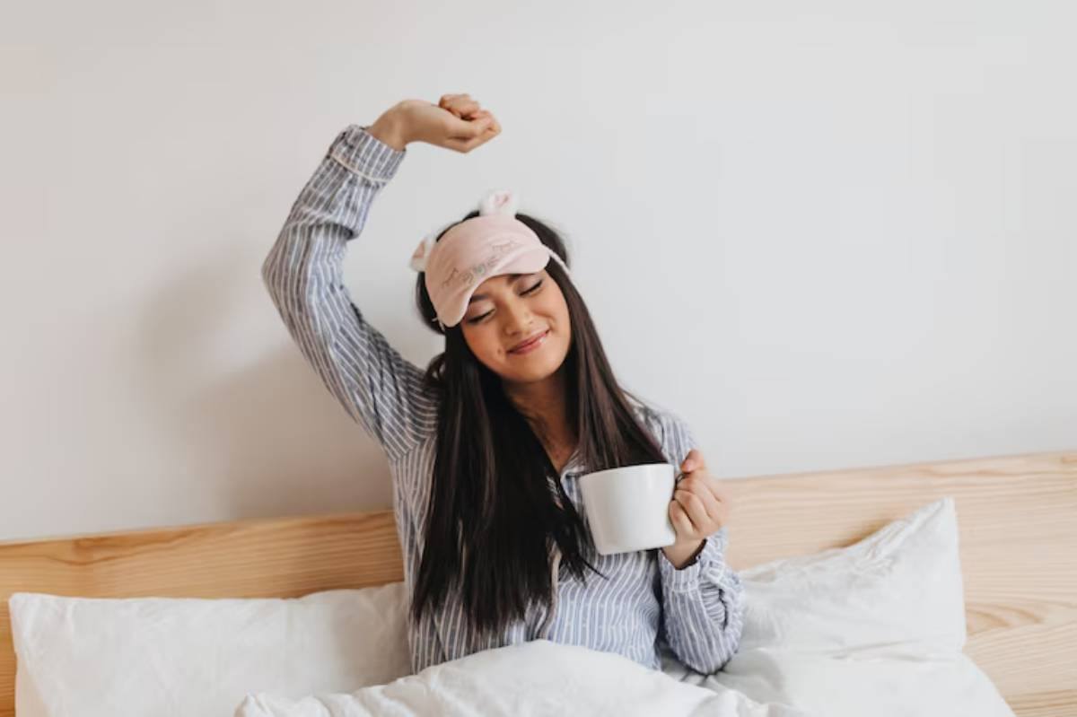 A woman in pajamas stretches while holding a coffee mug, seated on a bed surrounded by fluffy white pillows.